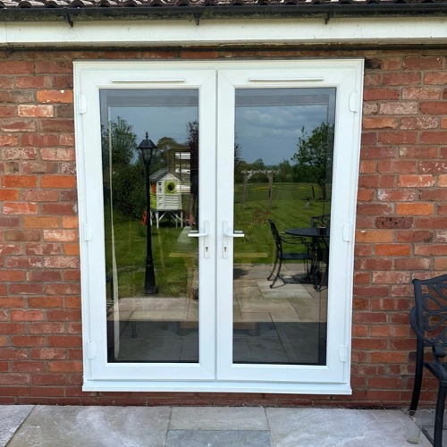 White UPVC French doors with glass panes set in a red brick wall, reflecting a garden with lawn, trees, and outdoor furniture.