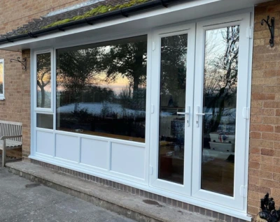 White UPVC French doors and large window panels installed on a brick wall reflecting trees and sky at sunset.