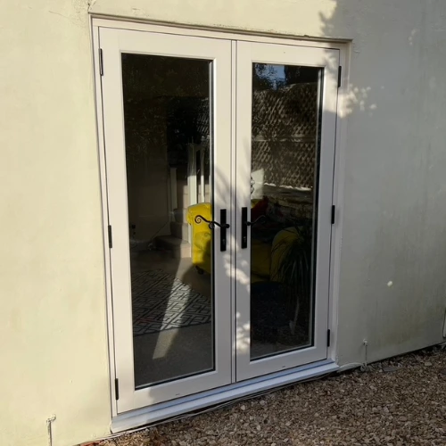 White uPVC French doors with glass panels installed in a beige exterior wall, reflecting outdoor surroundings.