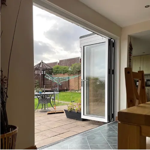 Open aluminium bifold door leading from a tiled indoor dining area to a paved backyard with garden furniture and plants under a cloudy sky.