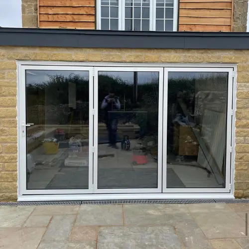 White aluminium bifold glass door installed in a stone and wood exterior wall, reflecting the outdoor patio and greenery.