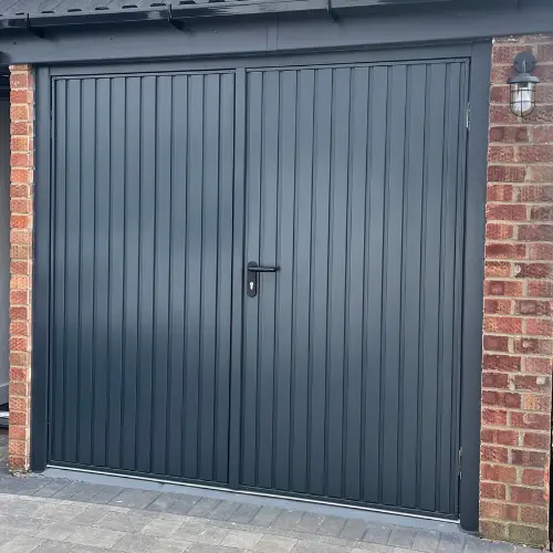 Black vertical panel garage door with central handle and lock, set in a brick wall with outdoor lamp on the right.