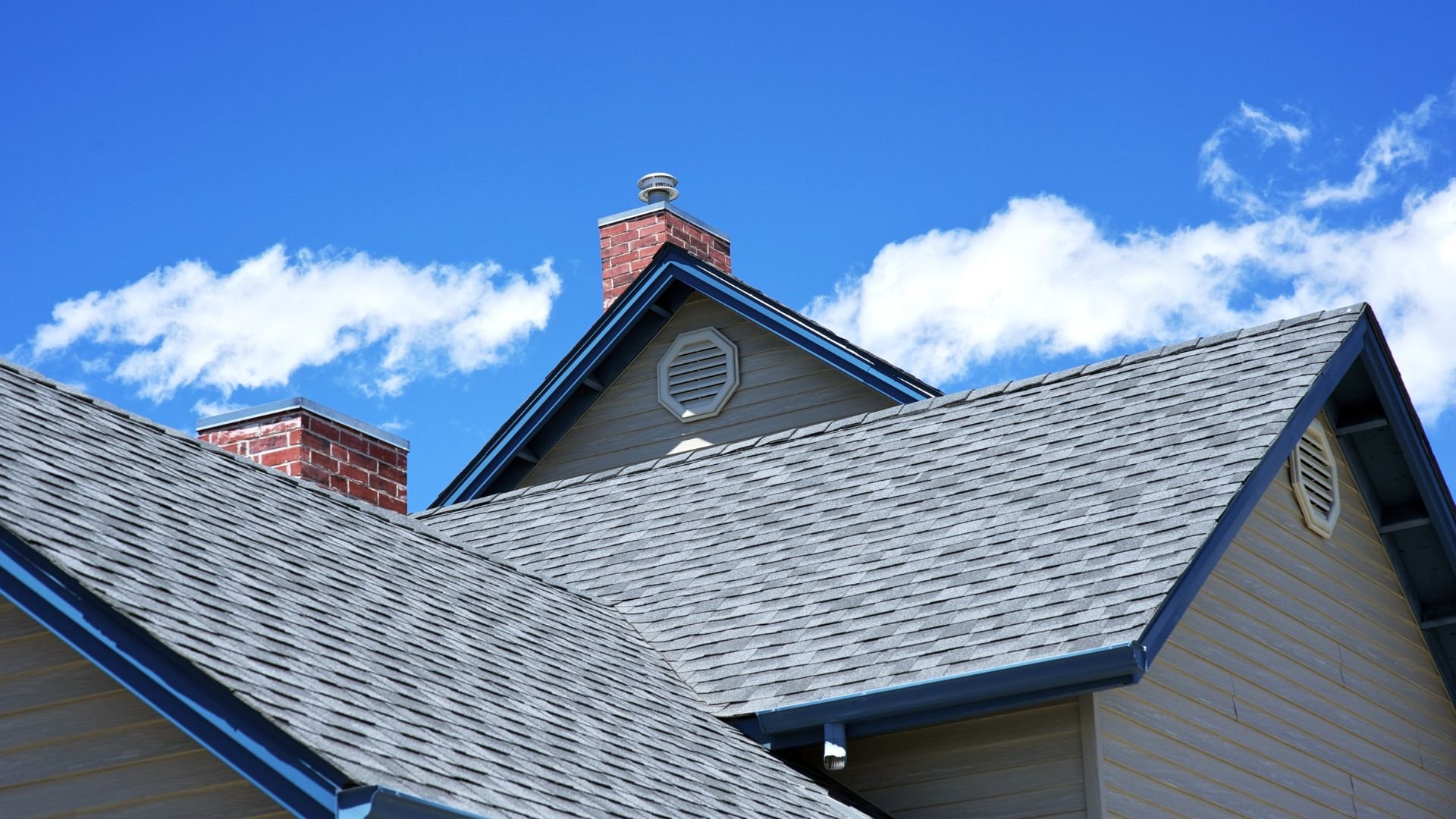 Gray shingle roof with brick chimneys against bright blue sky with clouds