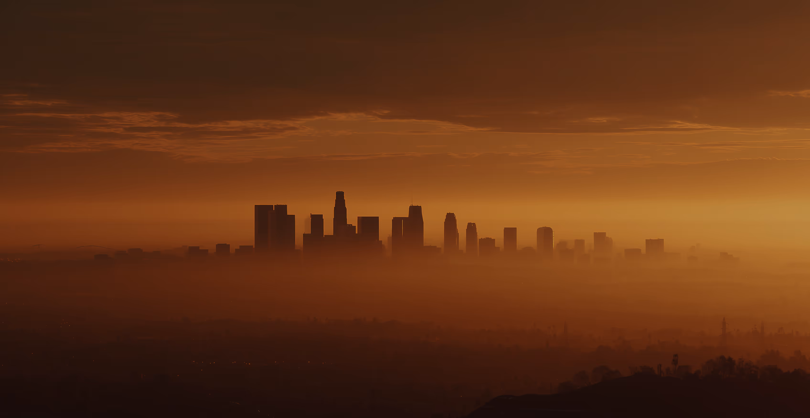 City skyline with tall skyscrapers fading into orange haze at sunset, with clouds above the horizon.
