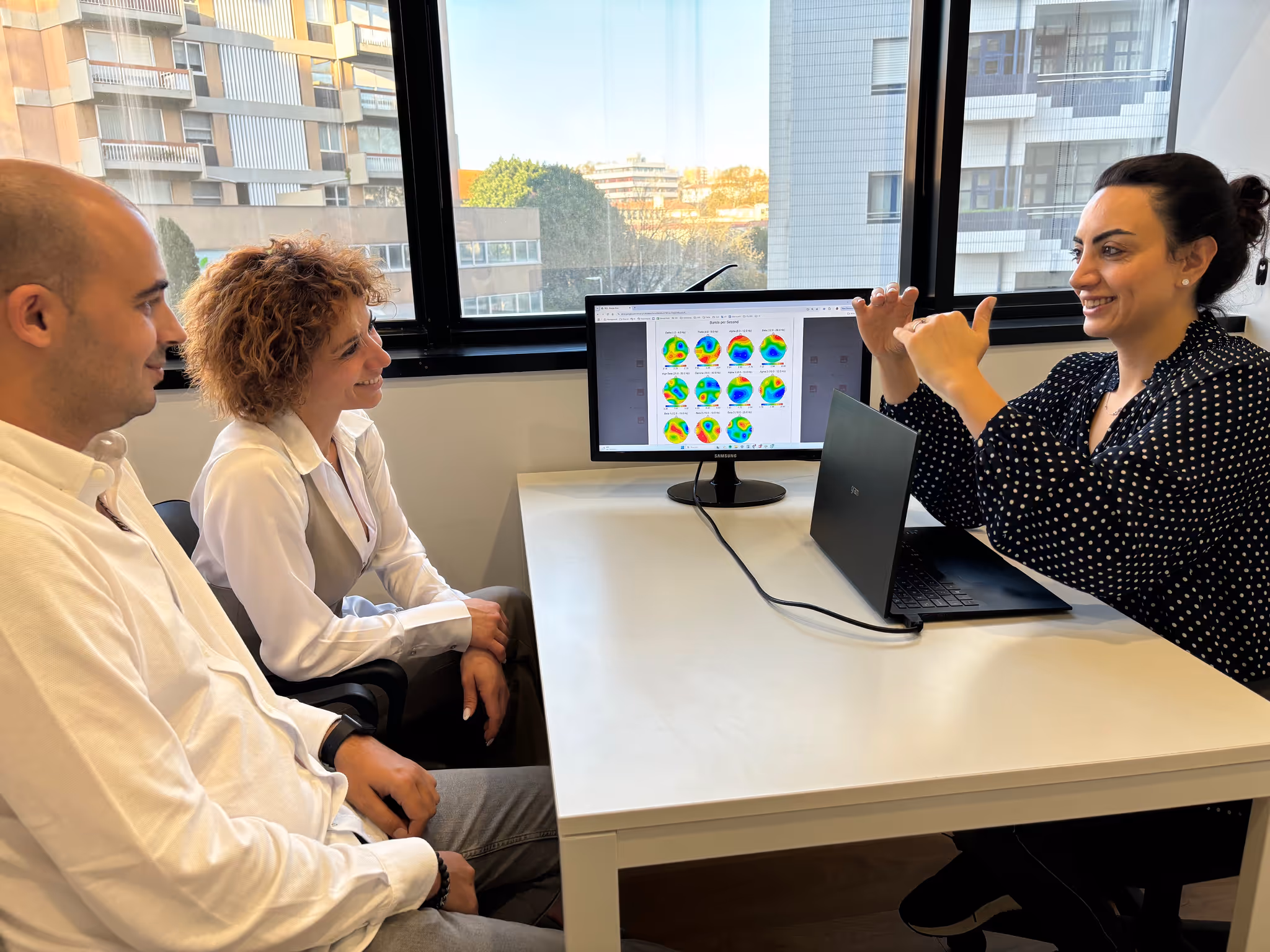 Profissional da NeuroImprove a explicar informação clínica a dois adultos, com apoio visual num ecrã, numa sala de consulta.