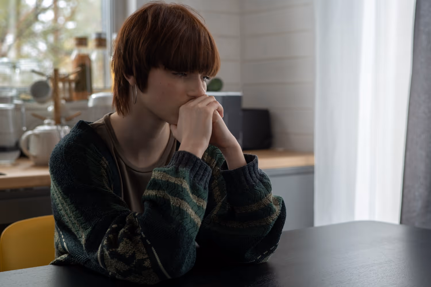 Adolescente sentada à mesa, com as mãos junto à boca, em atitude pensativa.