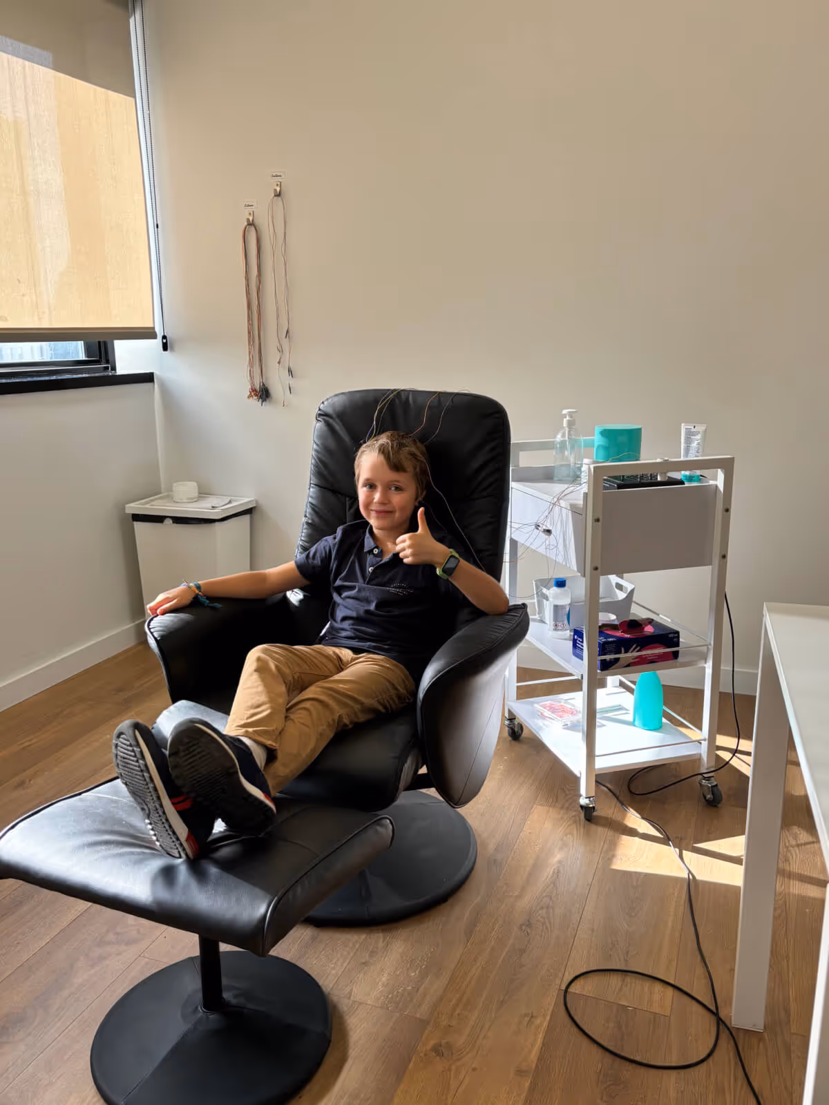 Criança sentada numa cadeira clínica durante uma sessão de neurofeedback, a sorrir e a fazer sinal positivo com o polegar.