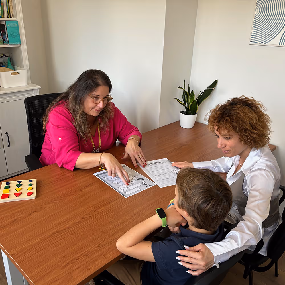 Criança acompanhada pela mãe e por uma profissional durante uma consulta de psicologia clínica, sentadas à mesa com material de apoio.