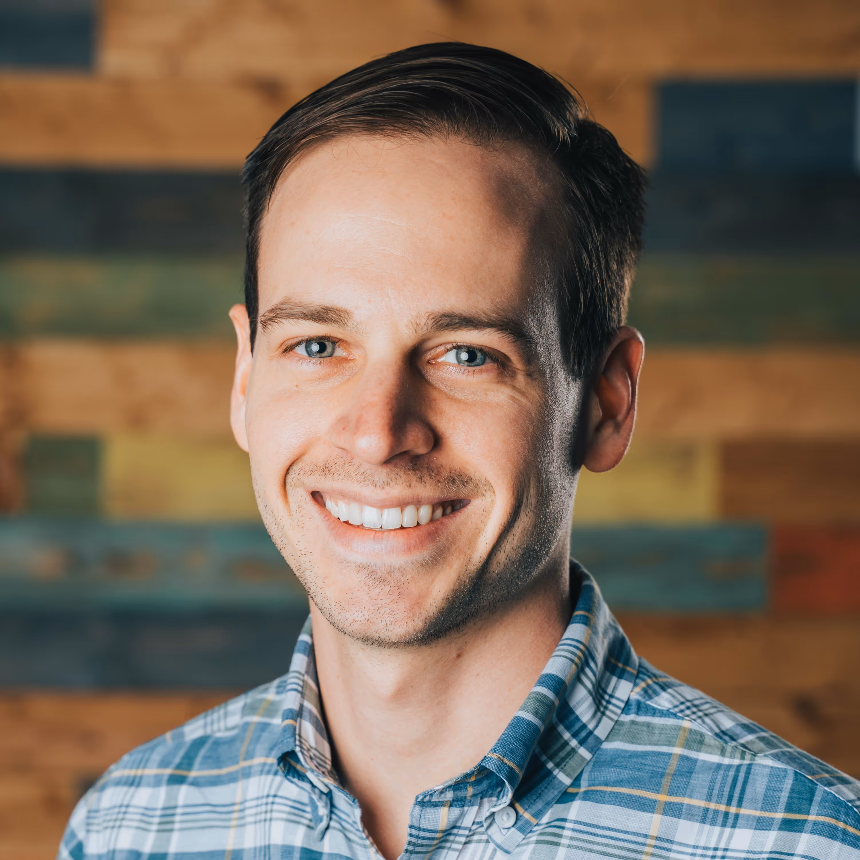Smiling man with short brown hair wearing a blue and green plaid shirt in front of a wooden textured background.