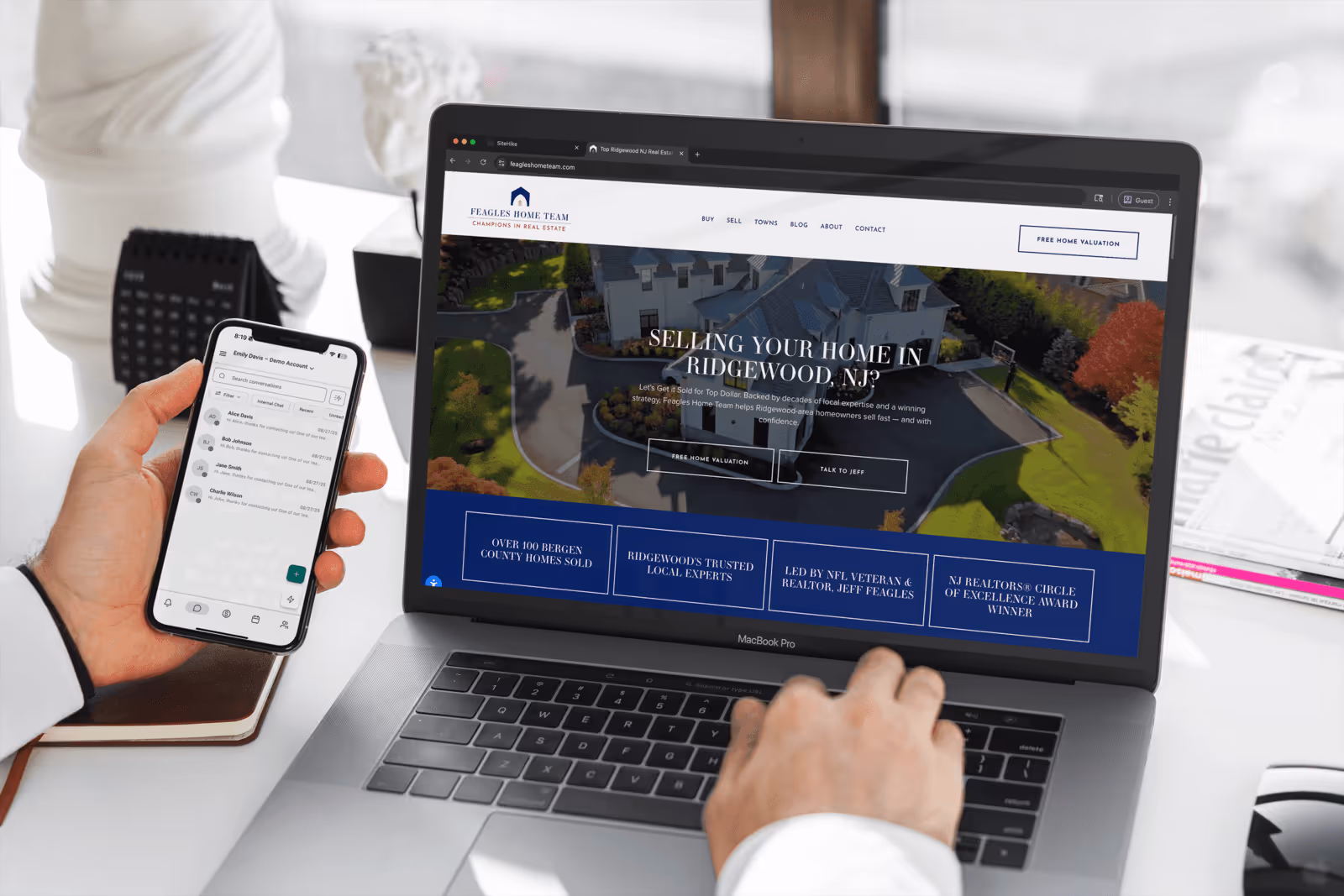 Person using a MacBook Pro displaying a real estate website for Ridgewood, NJ, while holding a smartphone showing a messaging app.