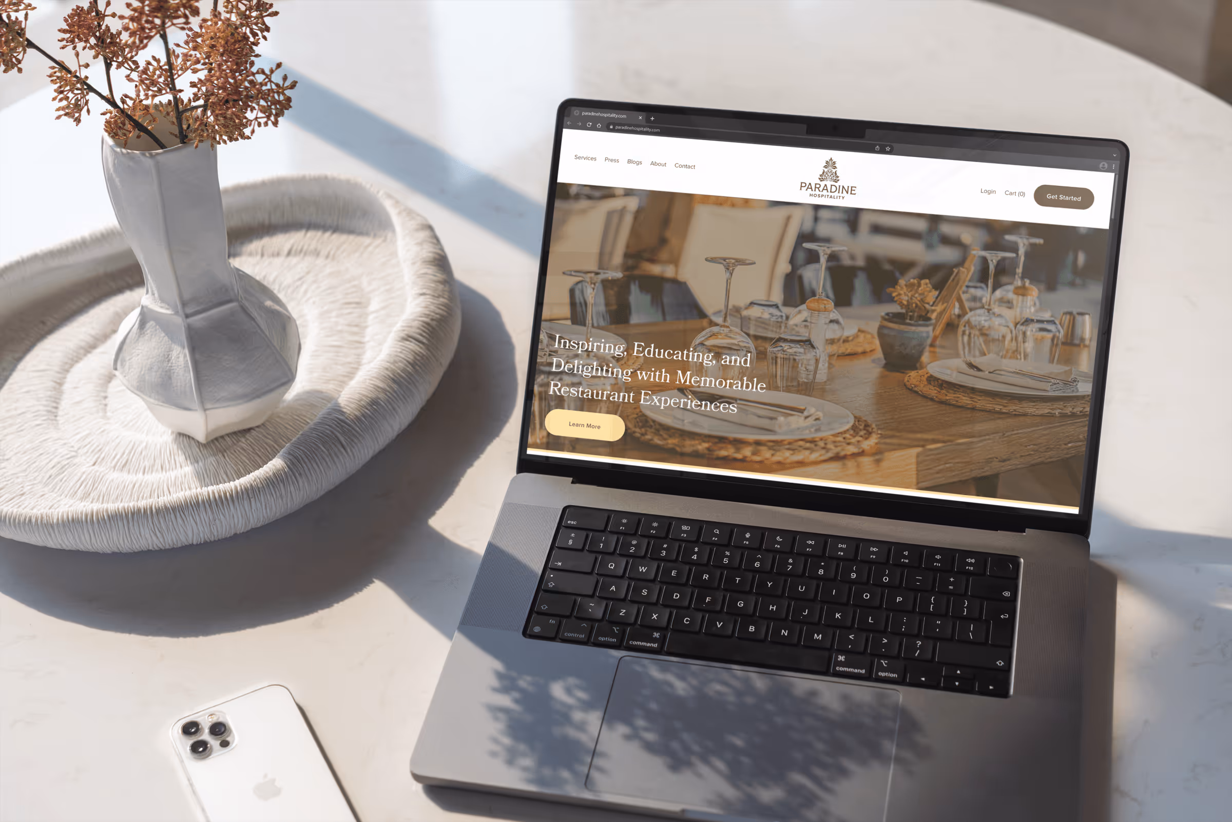 Laptop on a marble table displaying Paradine Hospitality website, next to a vase with dried flowers on a woven tray and a white iPhone.