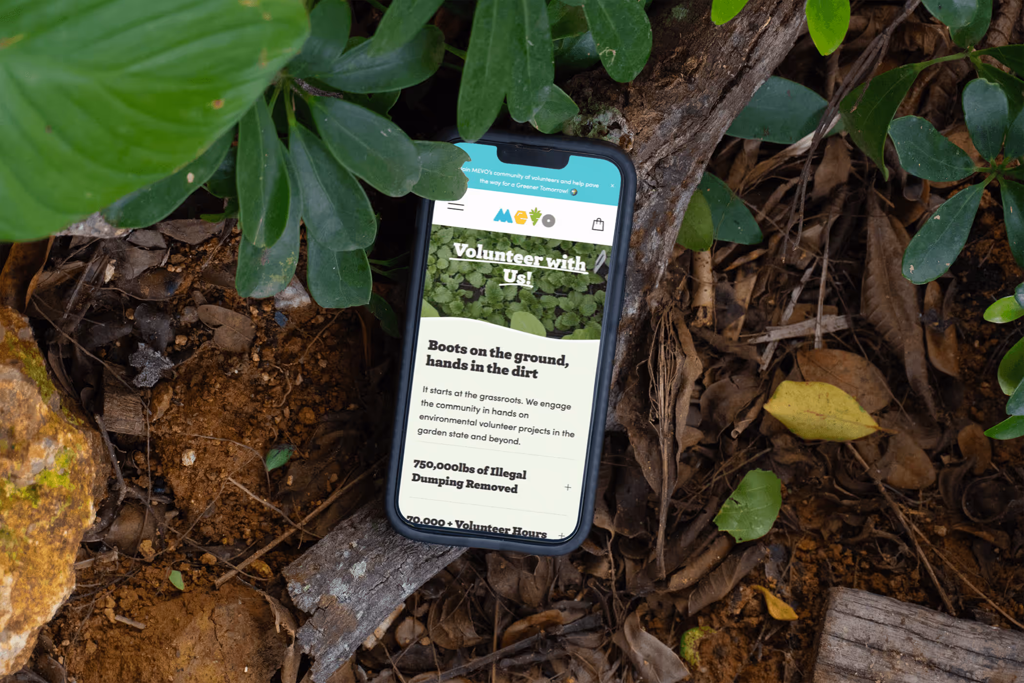 Smartphone displaying a volunteer recruitment webpage lies on soil surrounded by green leaves and plant debris.