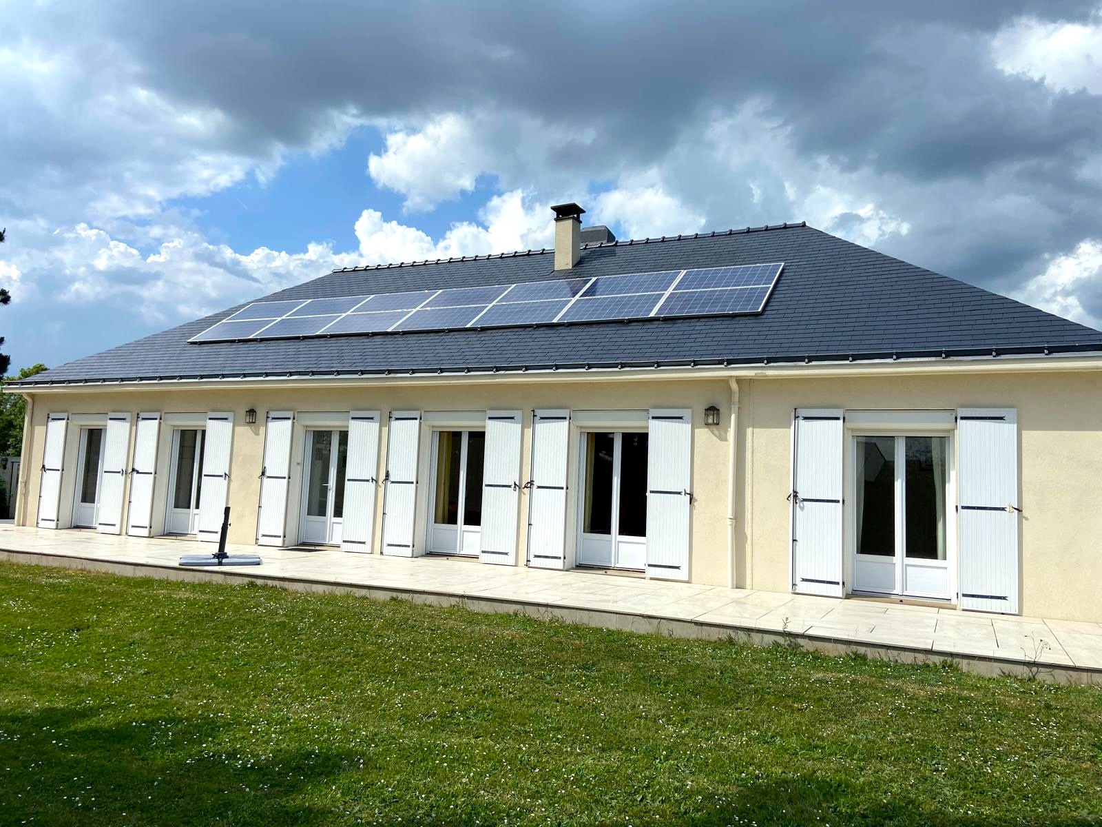 Maison beige avec fenêtres blanches et panneaux solaires sur le toit sous ciel partiellement nuageux.