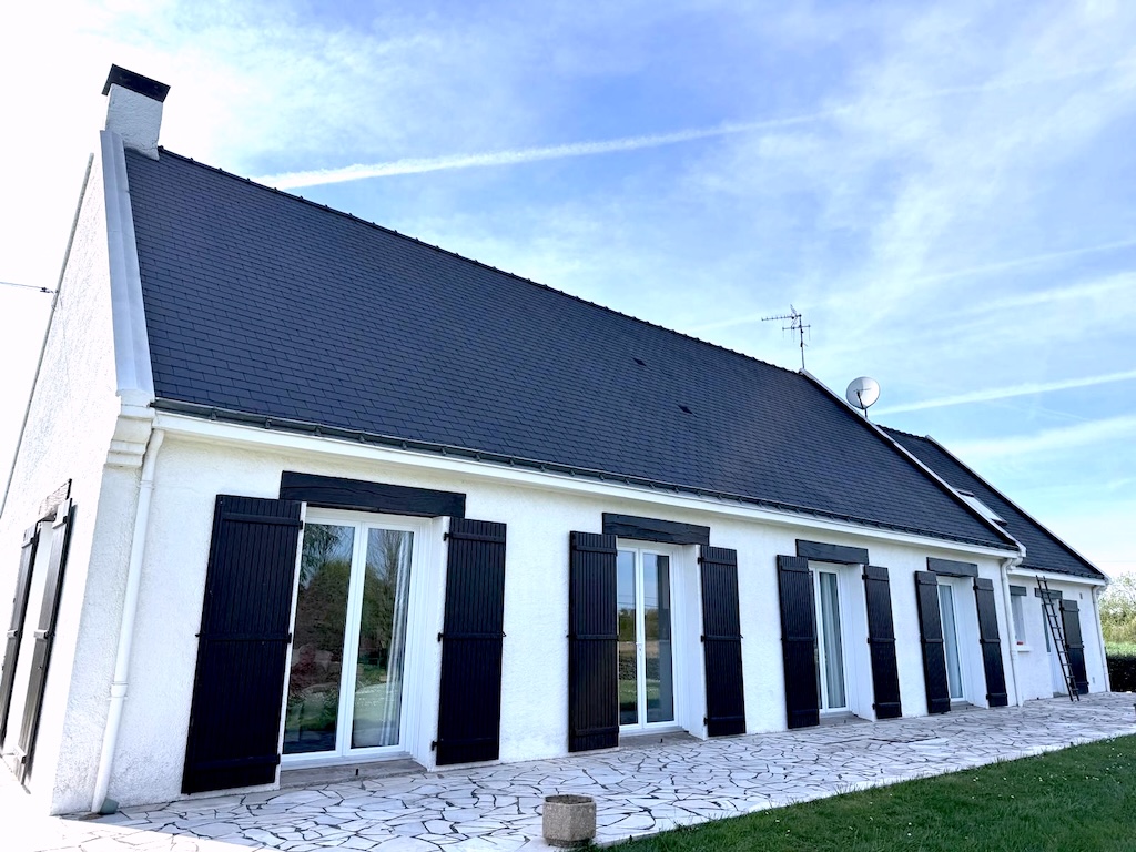Maison à toiture noire avec murs blancs, fenêtres avec volets noirs et terrasse en pierre.
