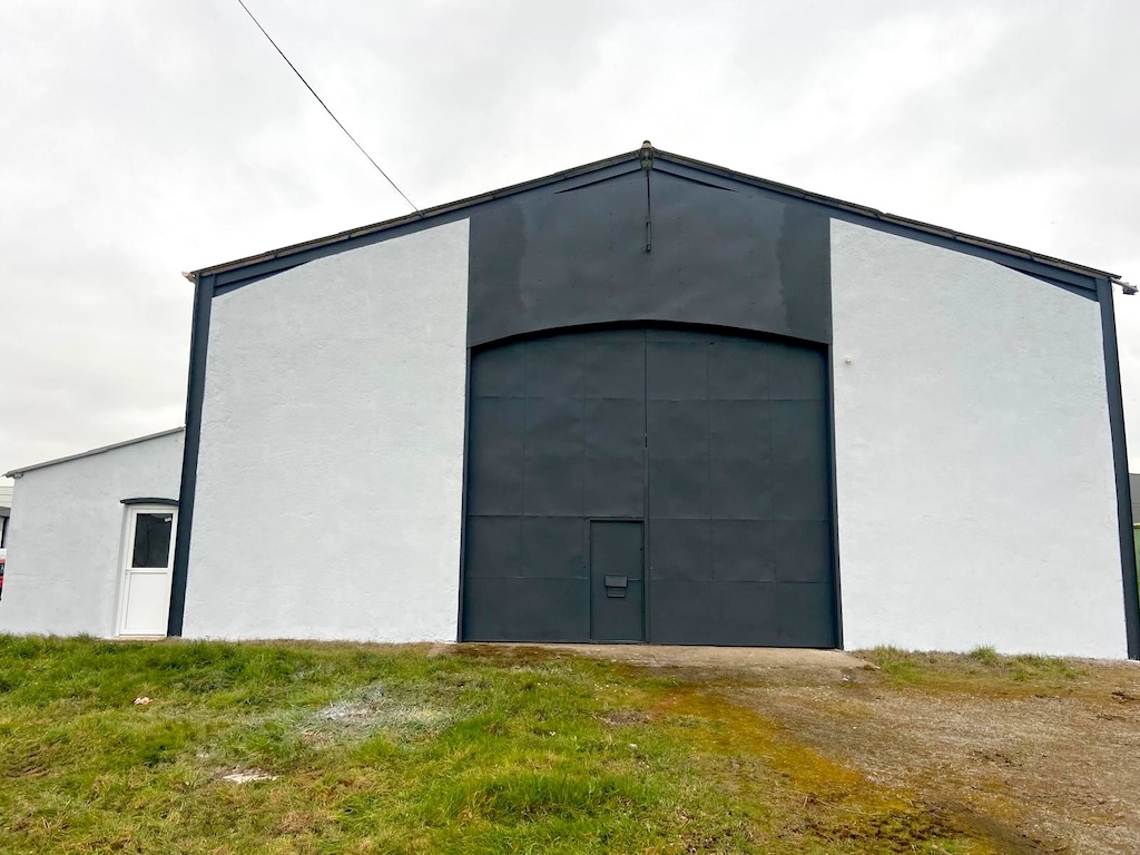 Grand bâtiment industriel blanc avec une grande porte noire au centre sur un terrain herbeux.