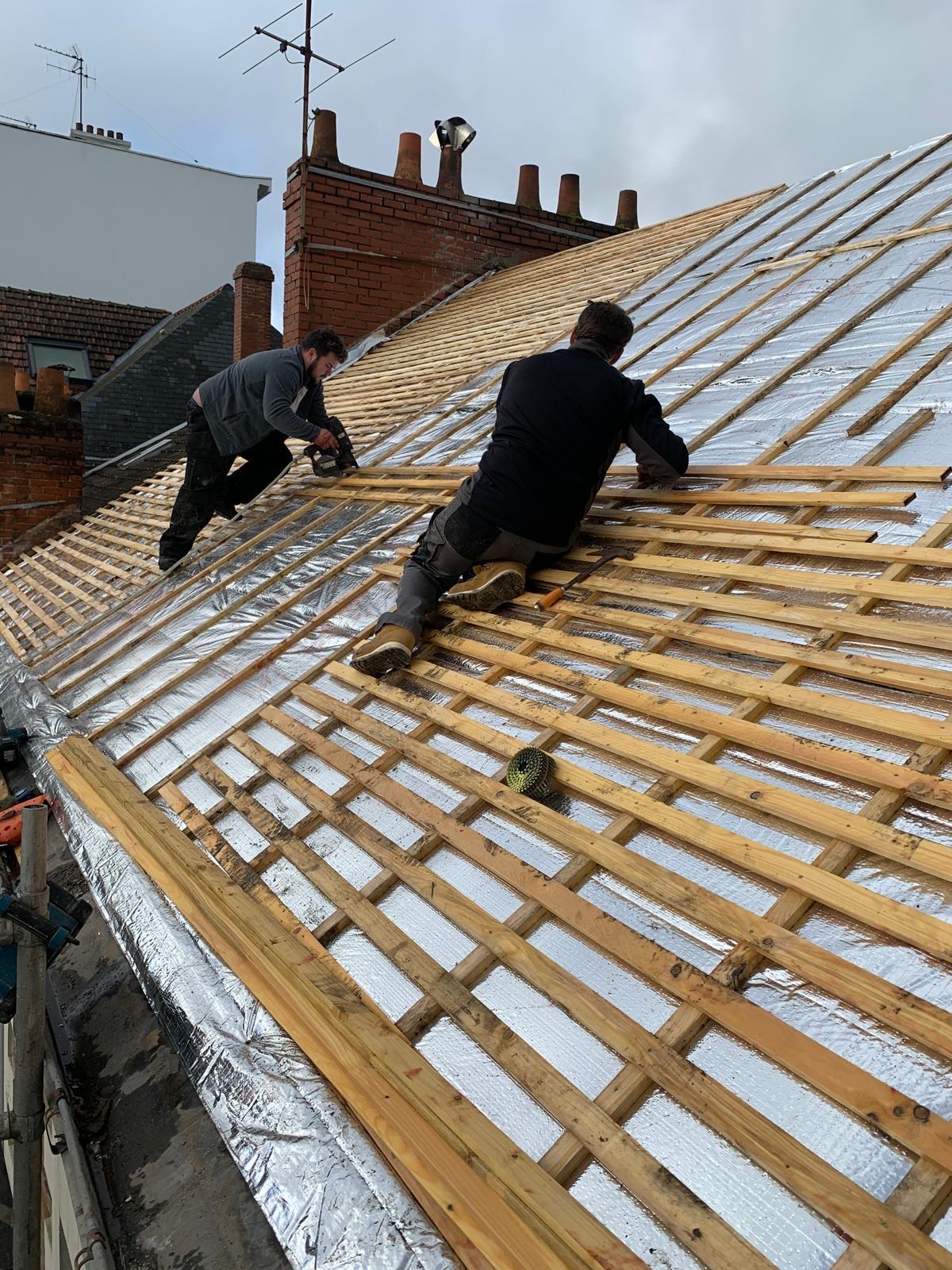 Deux ouvriers travaillent sur l'installation de liteaux en bois sur un toit incliné.