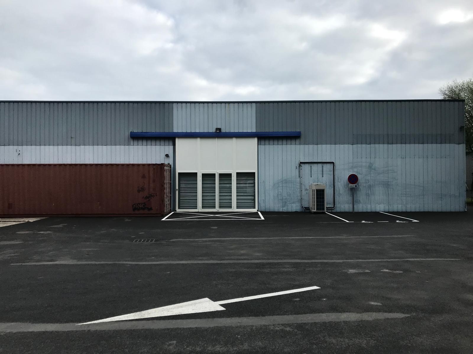 Façade d'un bâtiment industriel gris avec conteneur, porte blanche et panneau stationnement interdit.