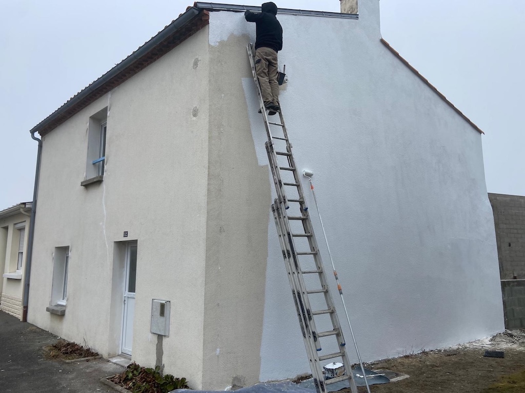 Un homme sur une échelle peint le mur extérieur d'une maison avec une peinture blanche fraîche.