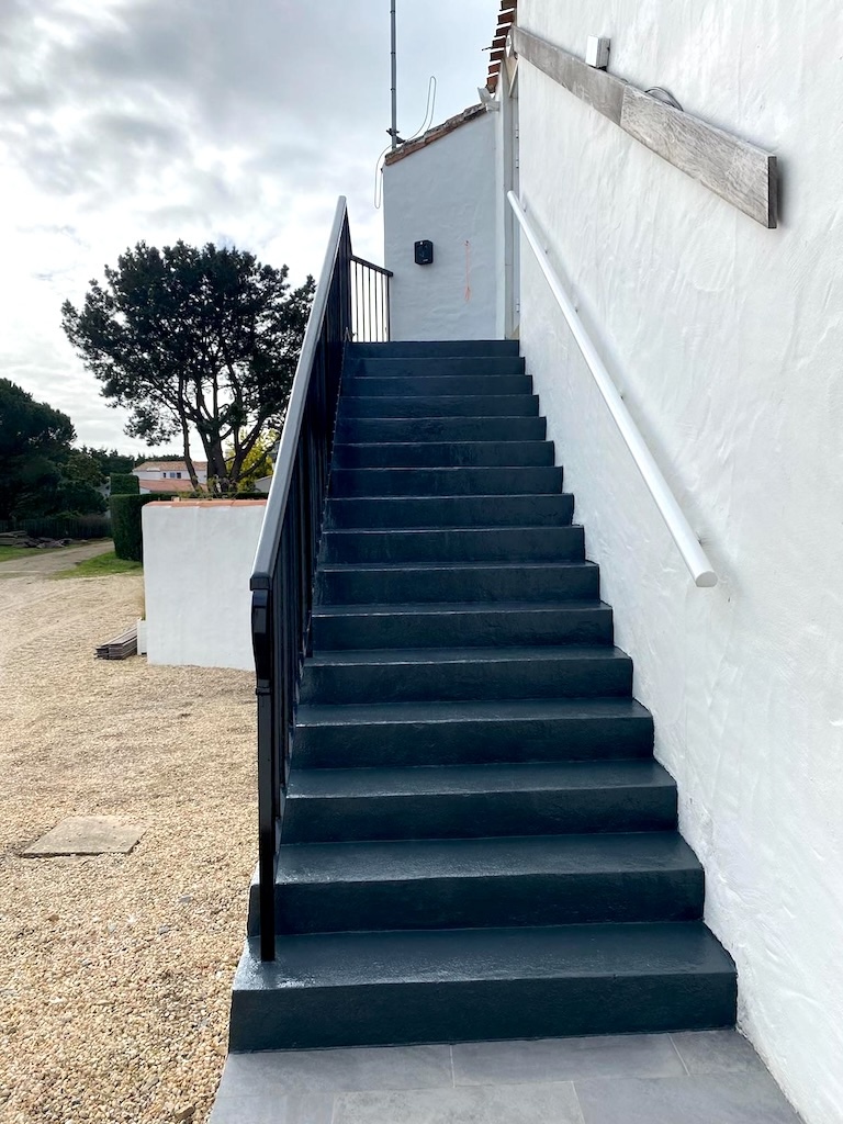Escalier extérieur en béton noir avec rambarde noire à gauche et mur blanc à droite.