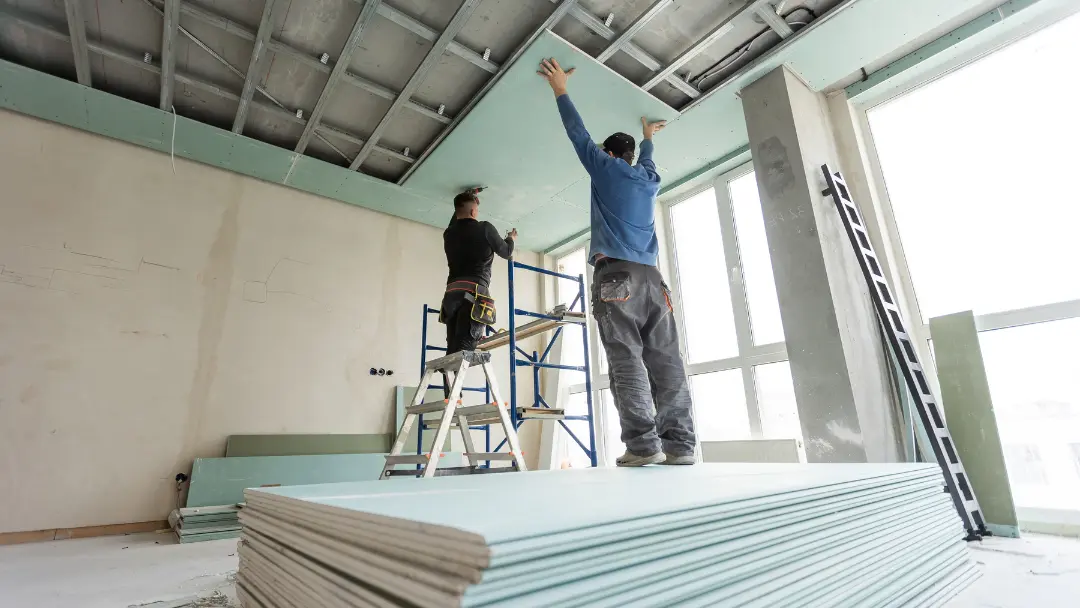 Two installers hanging drywall board on a ceiling grid with sheets stacked on the floor