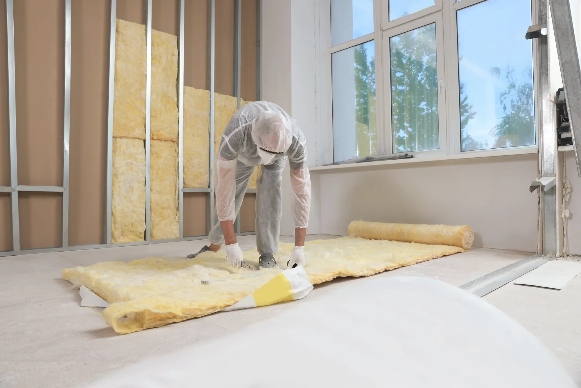 Installer in protective coveralls unrolling batt insulation beside a partly insulated stud wall
