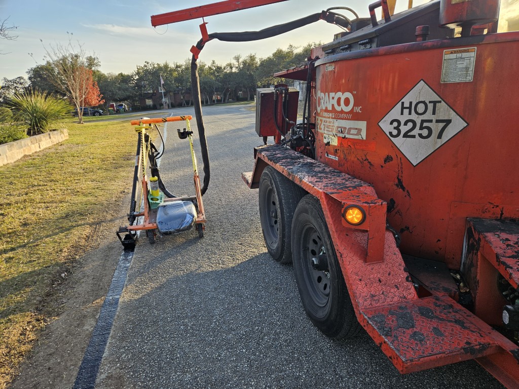 Red asphalt sealcoating machine with hose and handle on a road next to a freshly sealed black line.