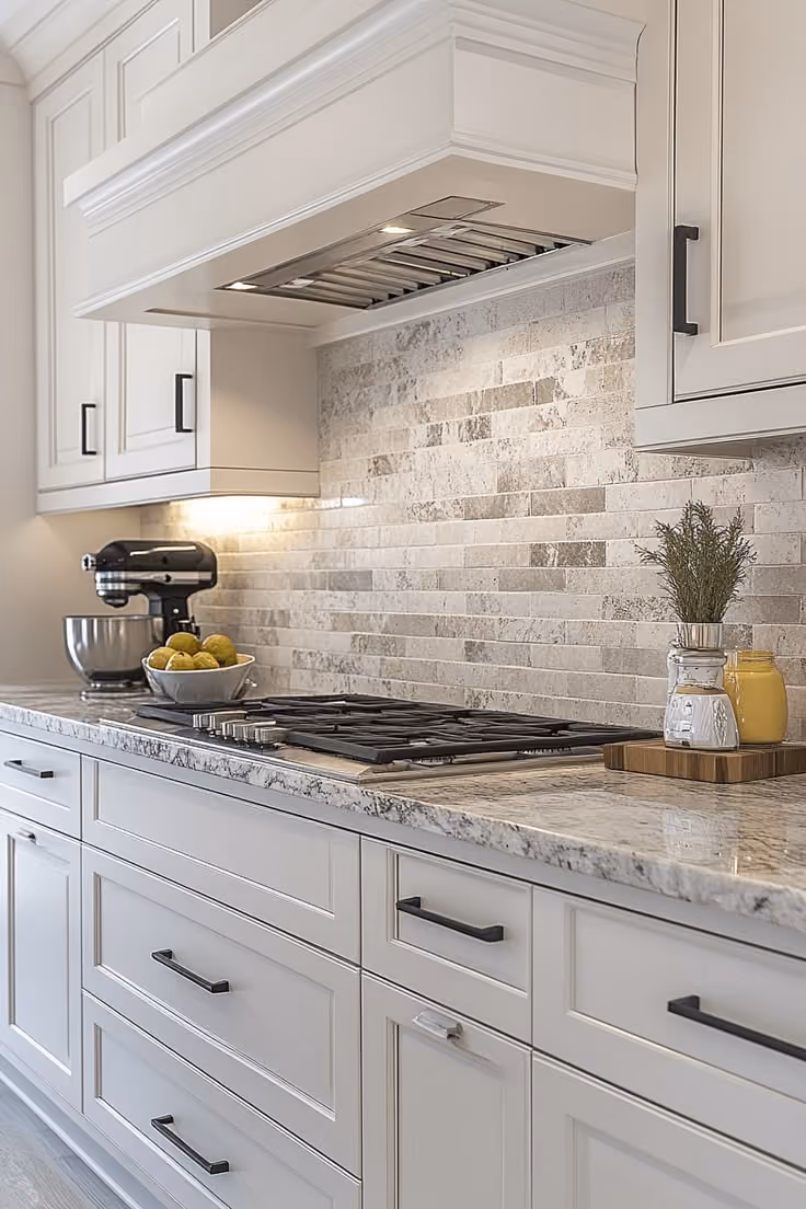 This may contain: a kitchen with white cabinets and marble counter tops, along with an oven hood over the stove