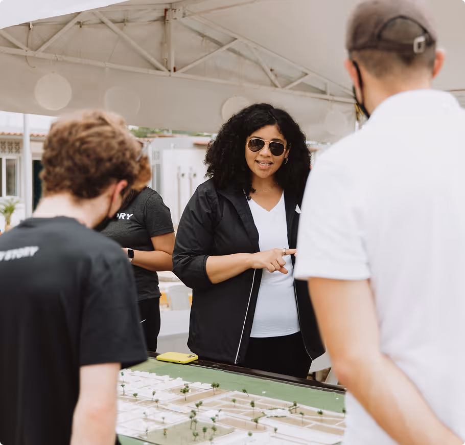 A woman wearing sunglasses and a black jacket speaks to a small group gathered around an architectural model during an outdoor meeting.