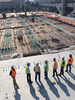 Group of construction workers in safety vests and helmets standing on a building site with visible rebar and piping