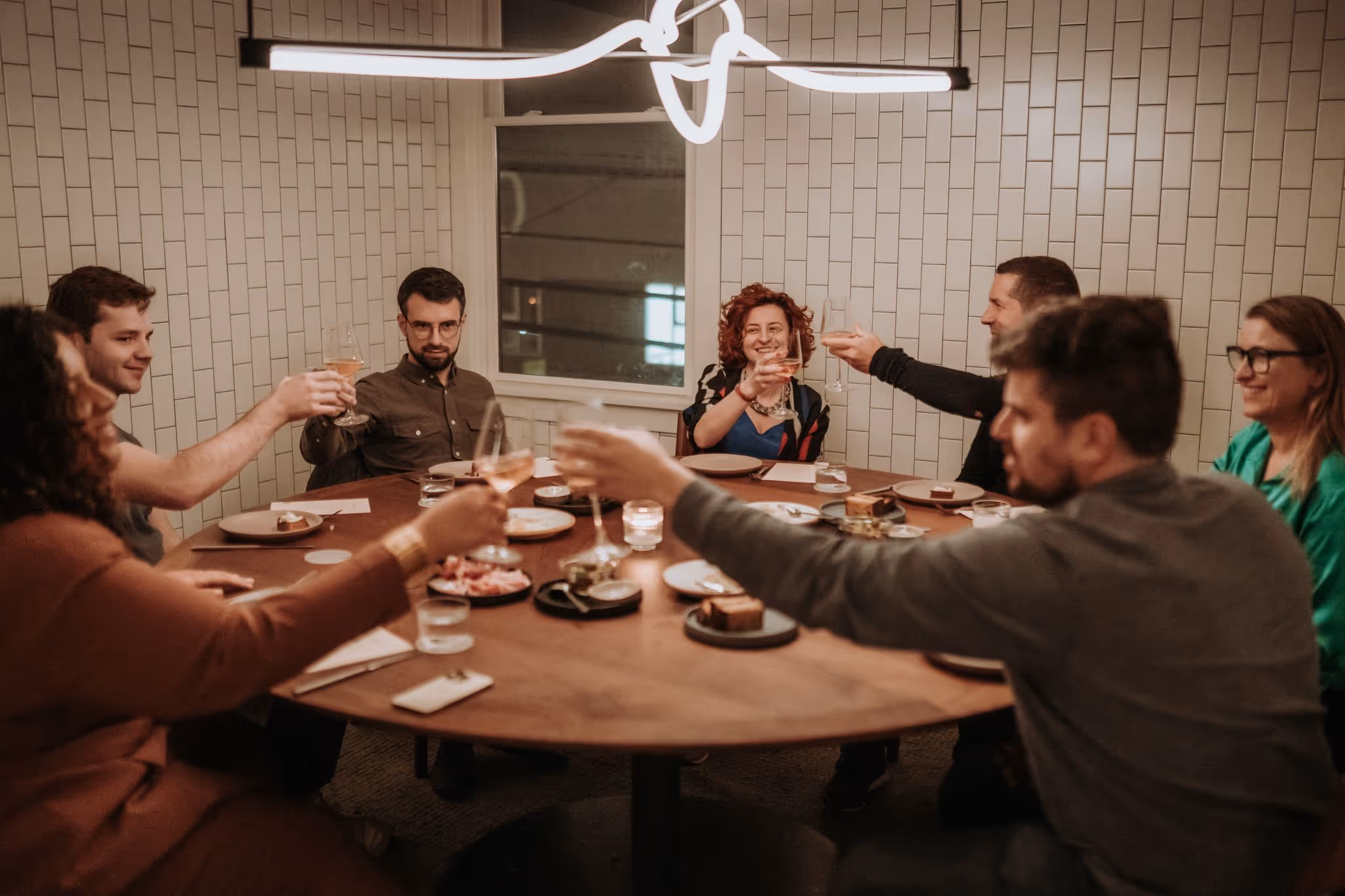 Group of people sitting around a dinner table raising glasses in a toast inside a warmly lit restaurant.