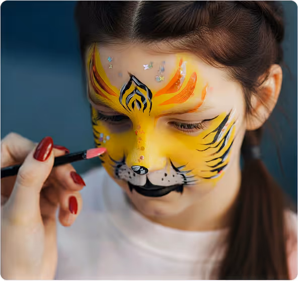 Niña con la cara pintada como un tigre amarillo con detalles negros y naranjas mientras alguien le aplica pintura rosa en la mejilla.