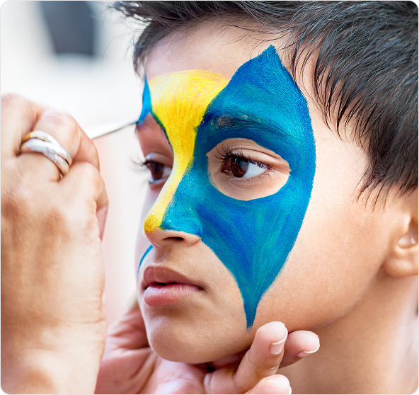 A girl with her face painted like a yellow tiger with black and orange details, while someone applies pink paint to her cheek.
