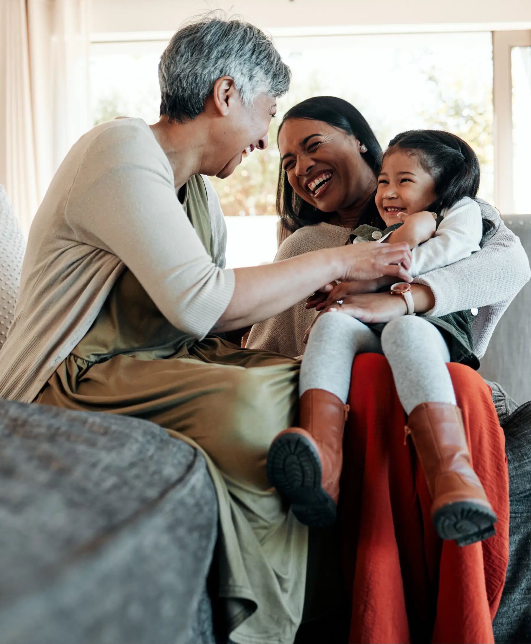 Grandmother, mother, and young daughter laughing together on a couch at home.
