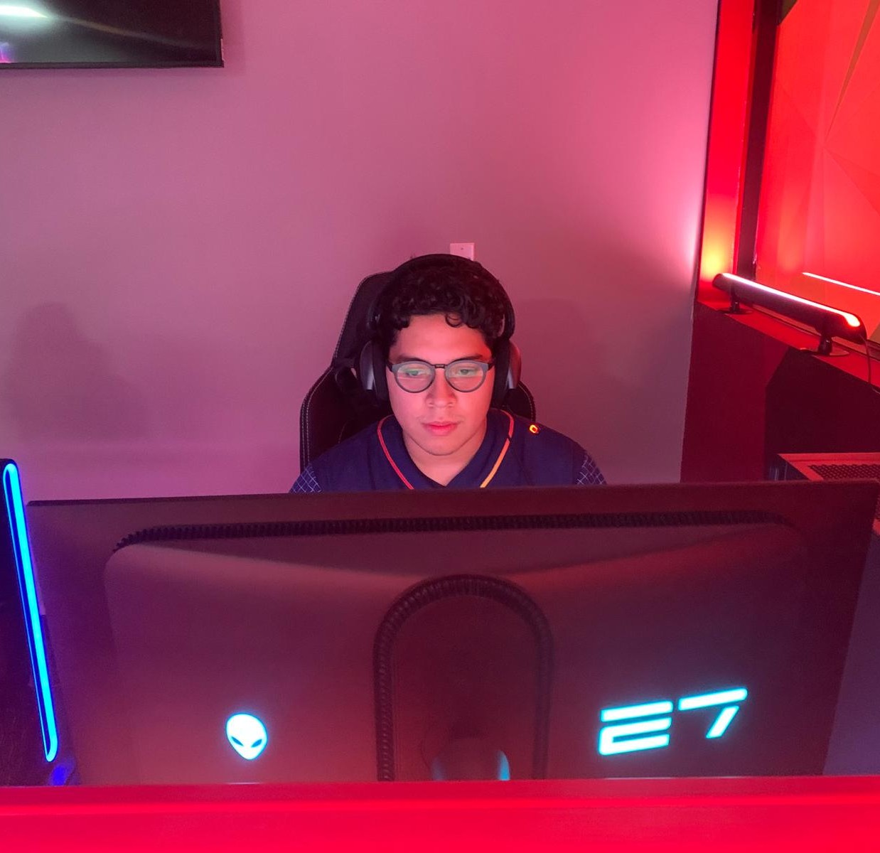 Man with glasses and headphones focused on a large Alienware 27-inch monitor in a dim room with red and purple lighting.
