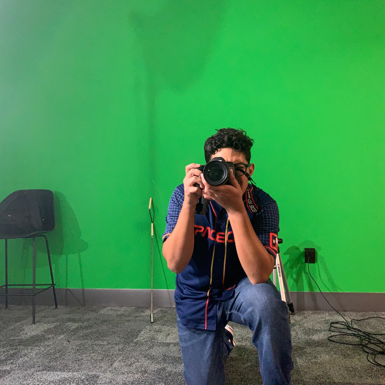 Person kneeling in front of a green wall, taking a photo with a DSLR camera.