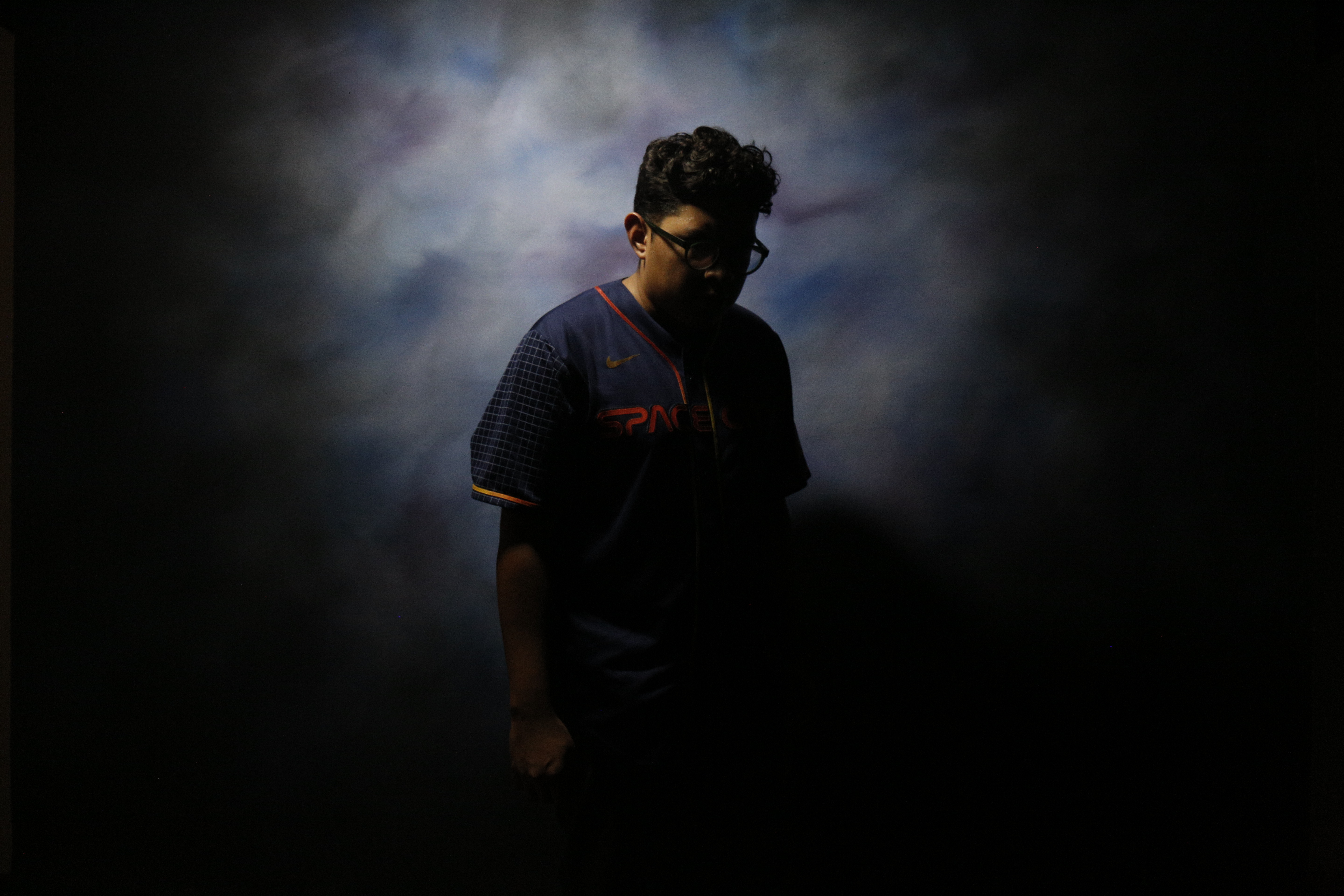 Person with glasses wearing a dark 'SpaceX' jersey standing against a cloudy blue and black background with face mostly in shadow.