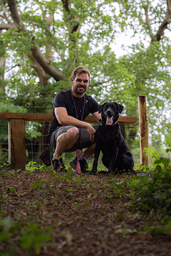 Man in black shirt crouching next to a black dog on a wooded path with green leaves above.