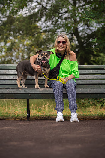 Woman in sunglasses and lime green shirt sitting on park bench with a happy dog on leash.