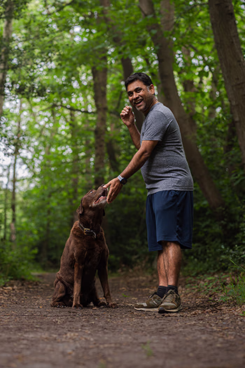 Man smiling and training a sitting brown dog on a forest trail surrounded by trees.