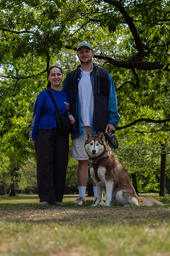 Man and woman standing in park with brown and white husky dog sitting in front of them.