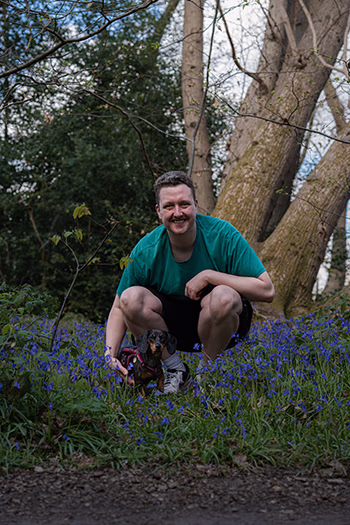 Man squatting in bluebell flowers in woods with small dog beside him on leash.
