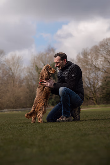 Man kneeling and facing a brown dog standing on grass in a park with cloudy sky and trees behind.