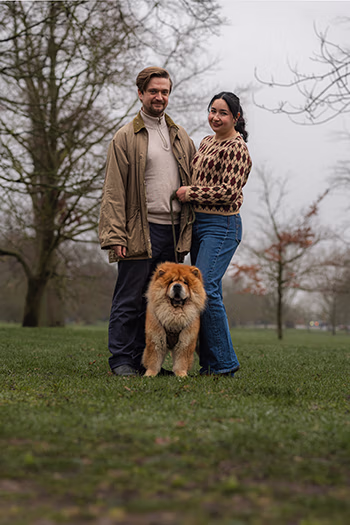 Smiling man and woman standing on grass with a fluffy dog between them in a park.