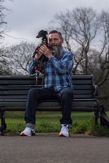 Man with a gray beard sitting on a park bench holding a small dog wearing a colorful sweater.