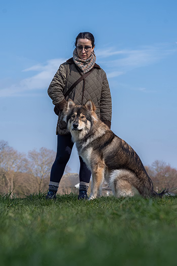Woman in a quilted jacket standing with a large dog sitting on grass under a blue sky.