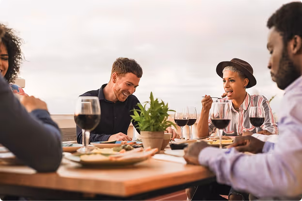 A diverse group of friends enjoys dinner at an outdoor table, laughing and engaging. Wine glasses and dishes are present, creating a joyful atmosphere.