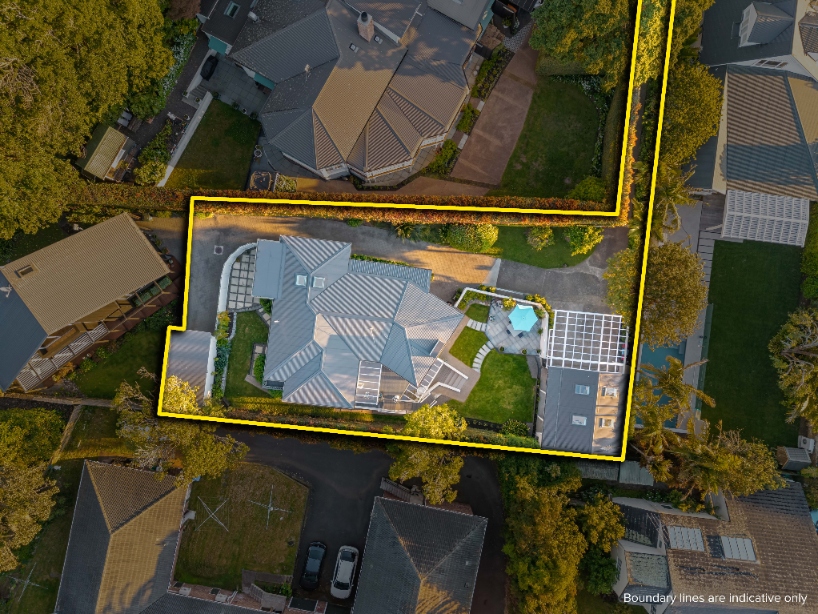 Aerial view of a residential property and large home with a complex grey roof and pool, outlined by a yellow indicative boundary line in a lush suburban setting.