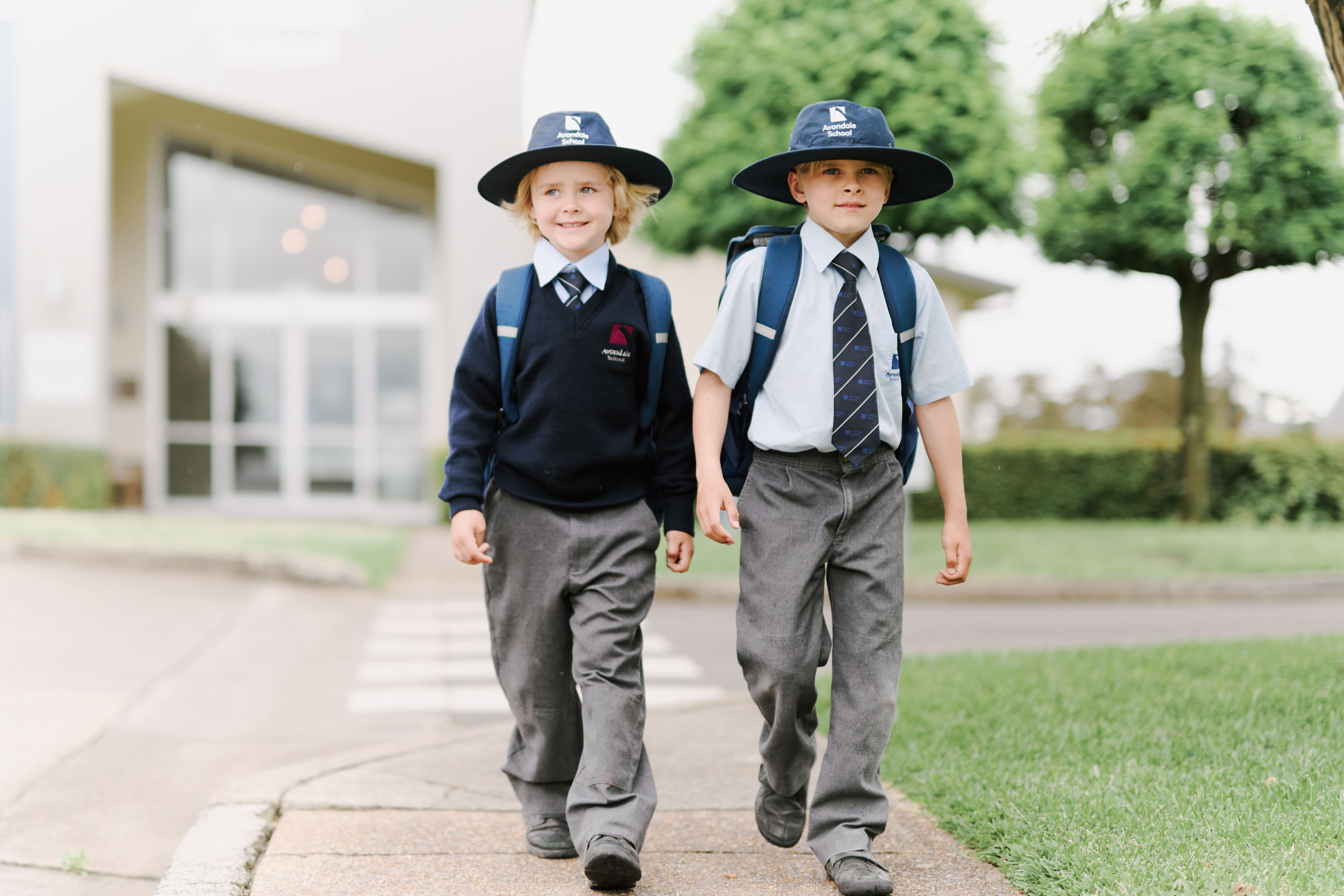 Two primary school children in uniform and wide-brim hats walk on a footpath, smiling and carrying backpacks, with a school building and greenery in the background.