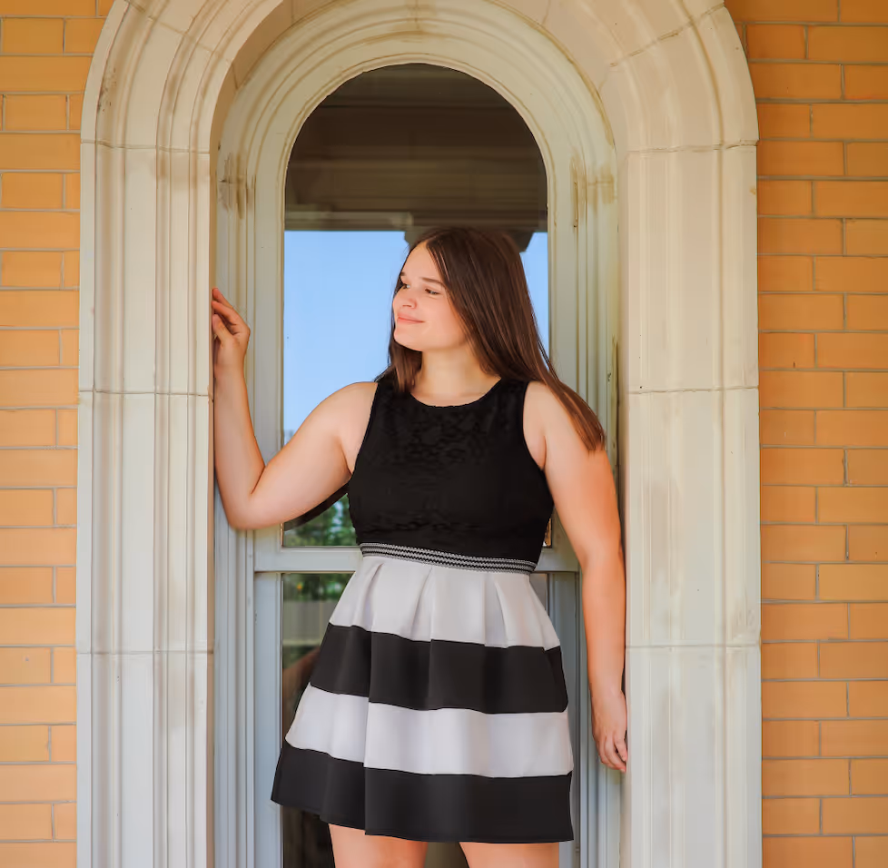 Young woman with long brown hair in a black and white striped dress standing in a decorative arched doorway.