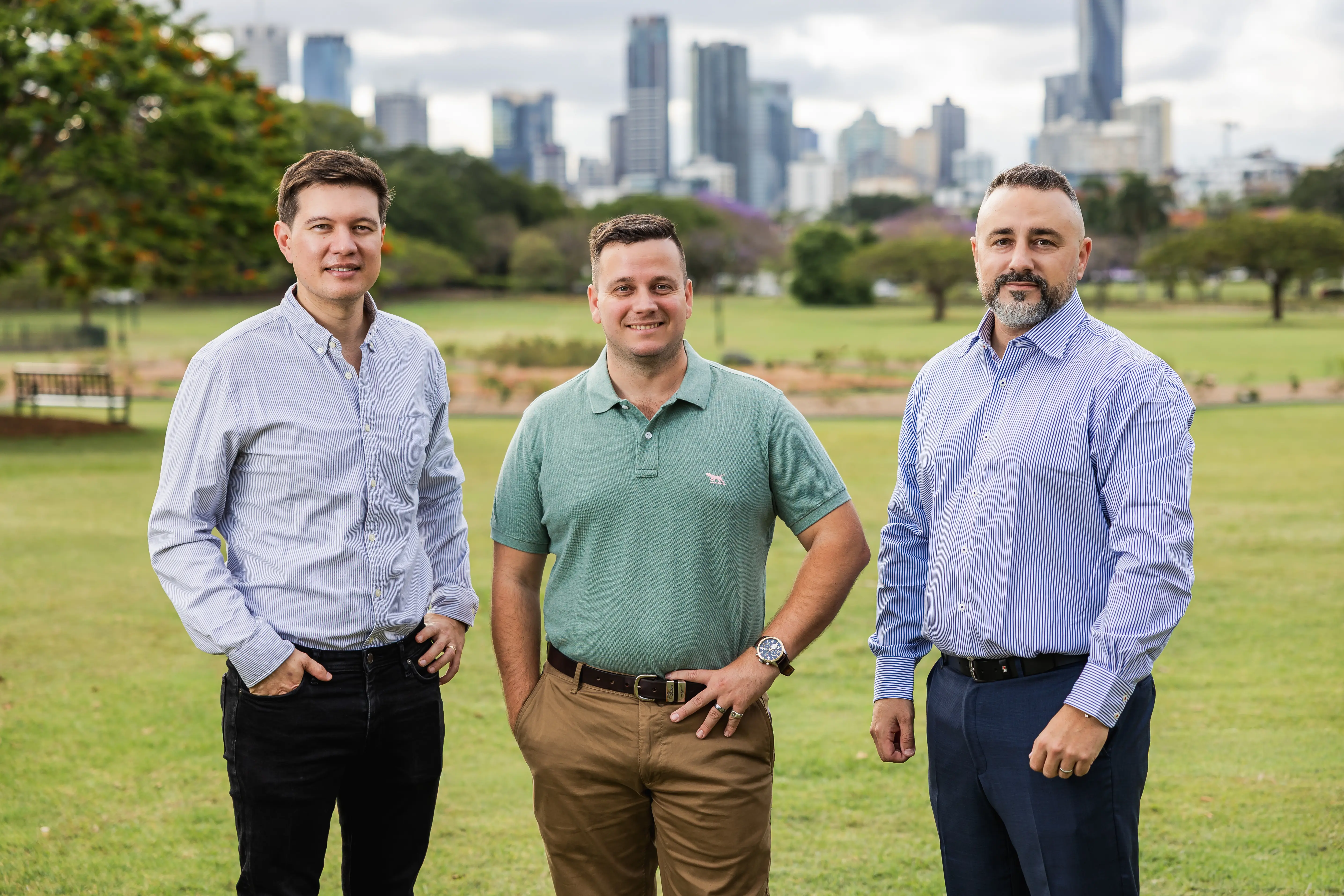 Three men standing on grass with an urban skyline and trees in the background.